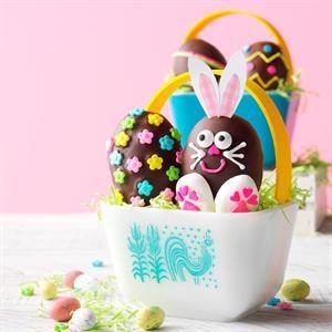 A white basket with blue rooster design contains colorful Easter eggs. One egg is decorated as a bunny with pink ears and whiskers. Nearby are small, multicolored candy eggs scattered on a wooden surface, set against a pink background.