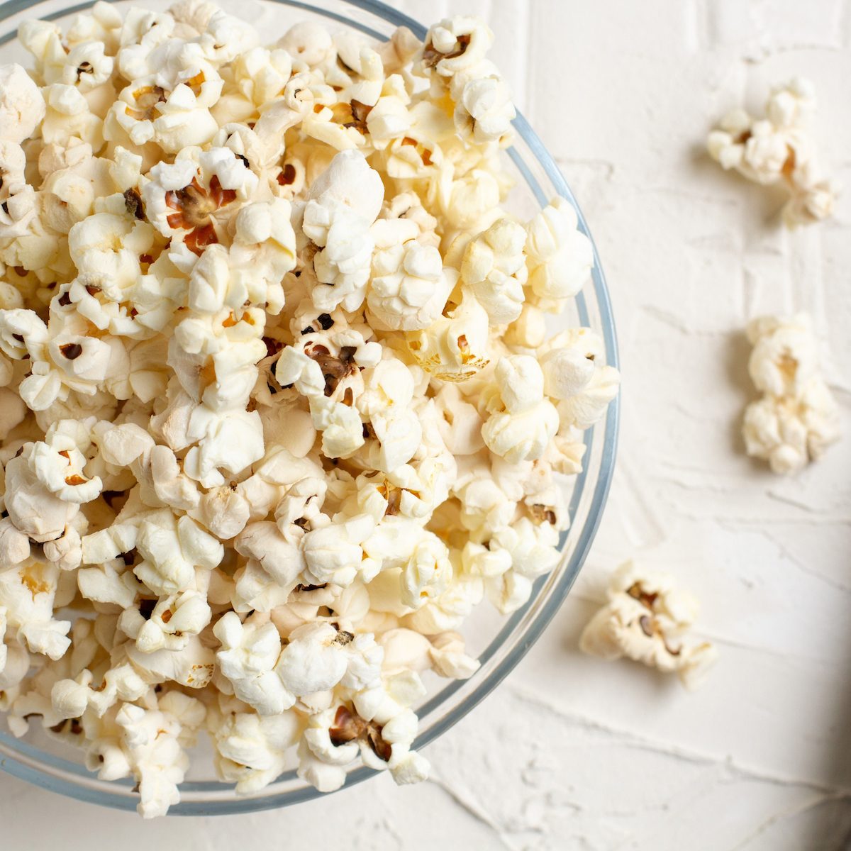 Popcorn in a transparent glass bowl on a white table with few pieces of popcorn beside the bowl.