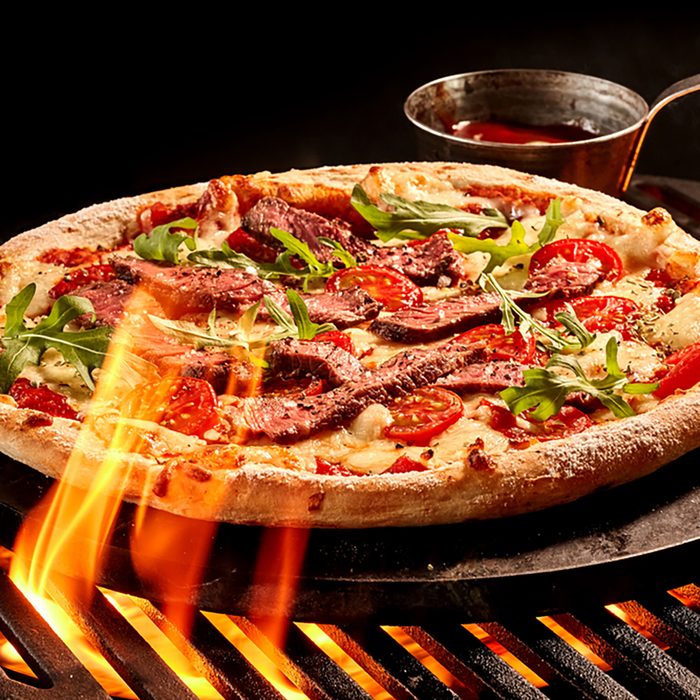 Flames reaching up toward meat and vegetable cheese pizza on grill. Hot sauce in bowl on iron plate