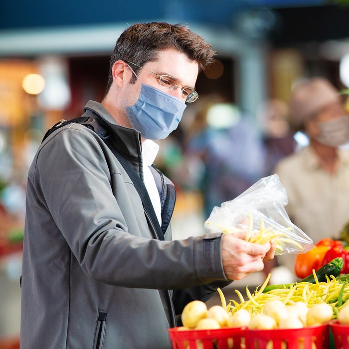 Young man wearing face cover selecting string beans at farmer's market to prevent spreading the covid 19 virus.