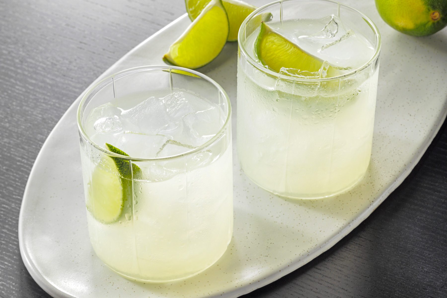 High angle view shot of Gimlet; served in ice-filled glasses; garnished with lime wedges; on white tray with lime wedges; black wooden base