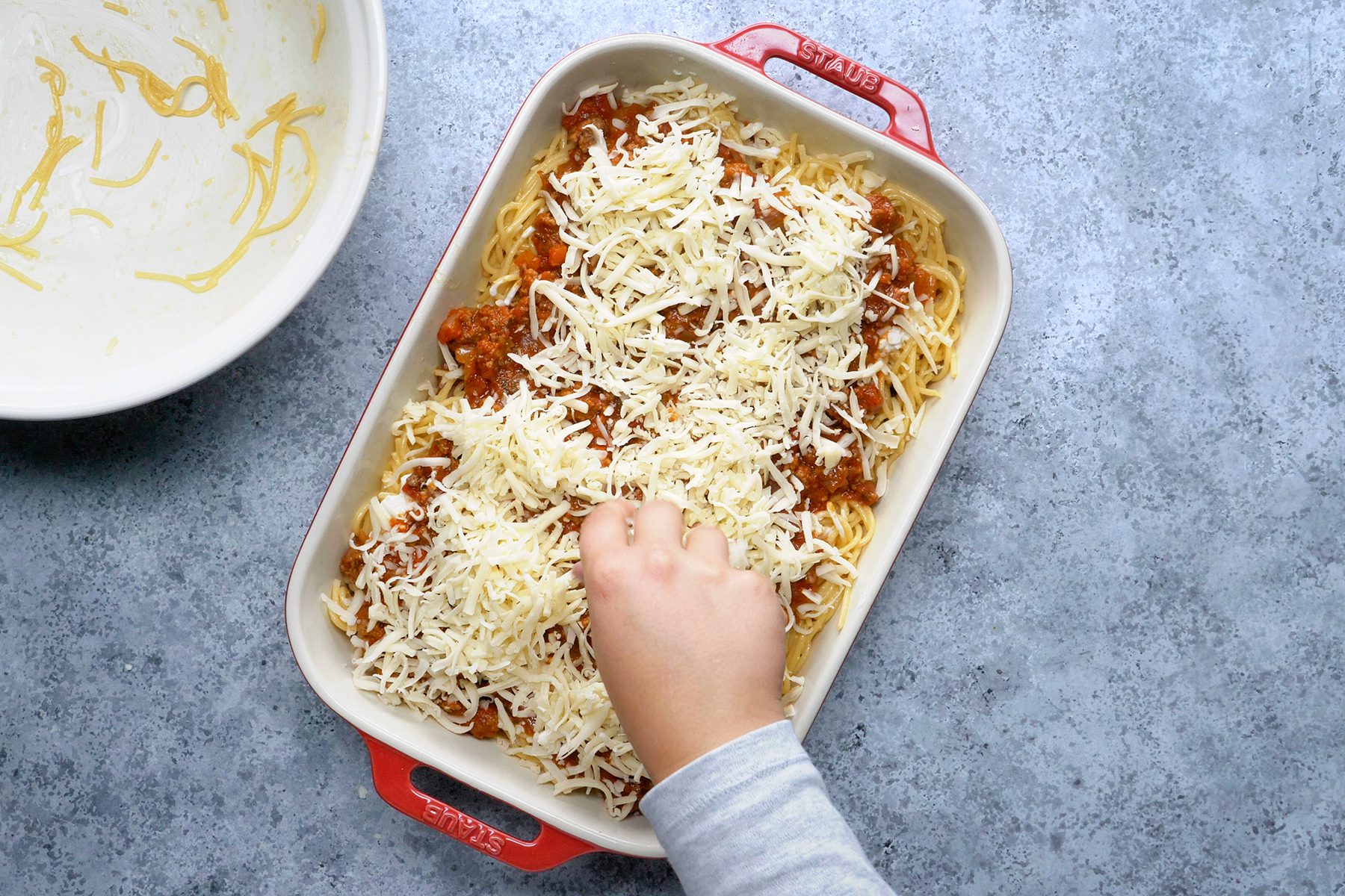 A child's hand sprinkles shredded cheese over a rectangular baking dish filled with spaghetti and meat sauce.