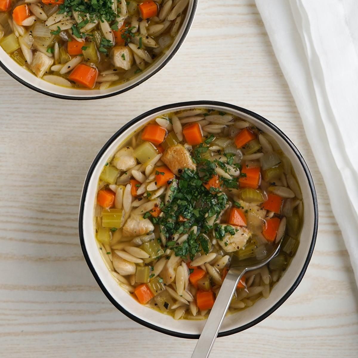 Two bowls of soup on a light wooden table. The soup contains chunks of chicken, diced carrots, celery, and orzo pasta, garnished with chopped herbs. A spoon rests in one of the bowls. A white napkin is placed nearby.