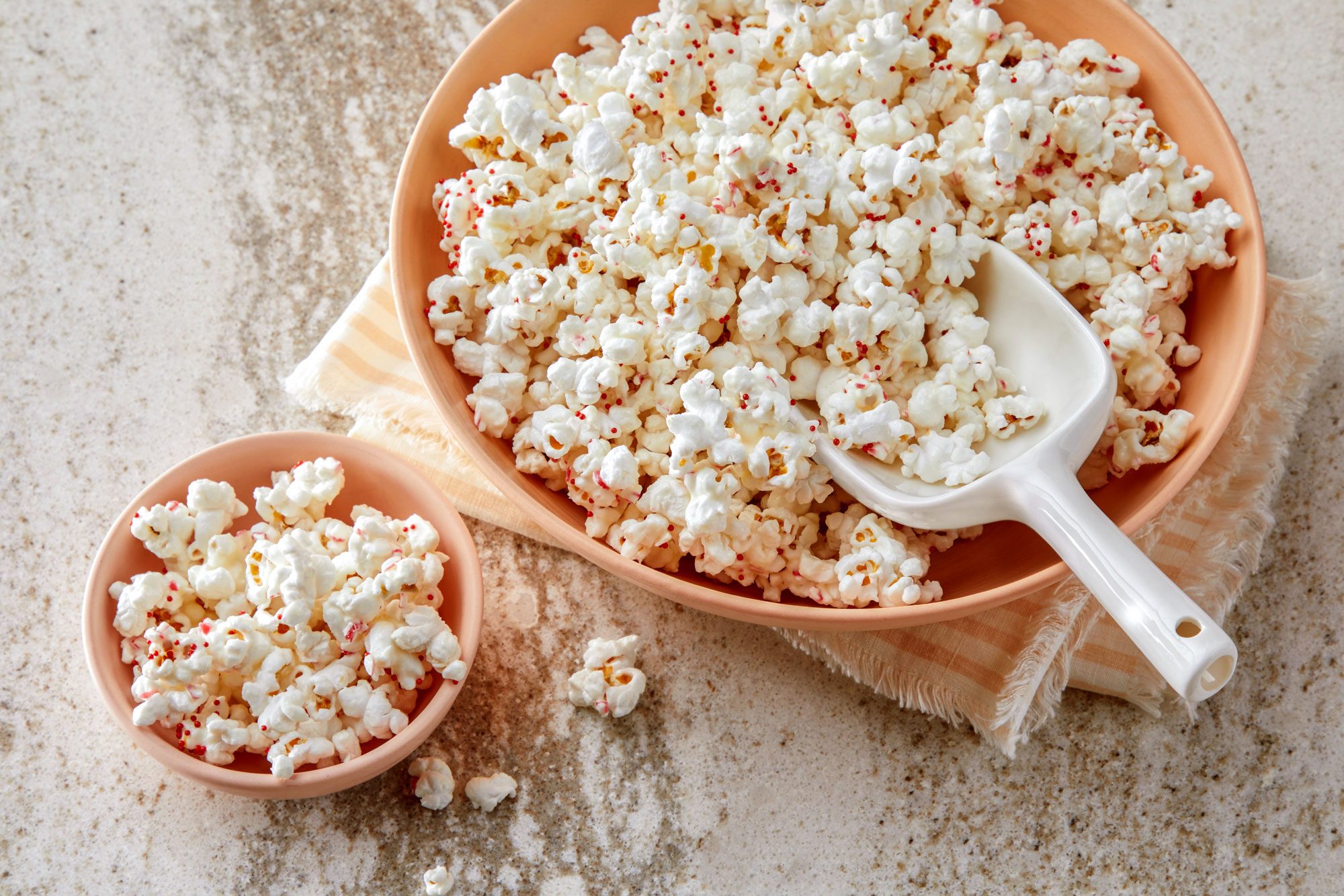 3/4th shot of two bowls of white popcorn with red sprinkles; a large bowl and a smaller bowl, both bowls are on a light colored surface with a piece of fabric underneath the larger bowl and a white serving spoon is next to the larger bowl, a few pieces of popcorn are scattered around the bowls on the surface