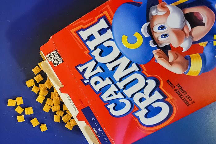 An open Cap'n Crunch cereal box lies on its side, spilling square cereal pieces onto a blue surface. The box features an illustrated sea captain in a blue uniform on the cover.