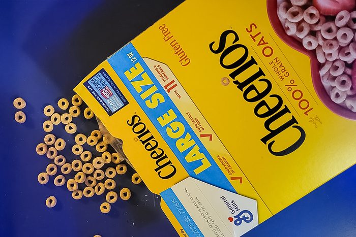 A large box of Cheerios cereal is tipped over, spilling circular cereal pieces onto a blue surface. The packaging highlights that it's gluten-free and made with 100% whole grain oats.