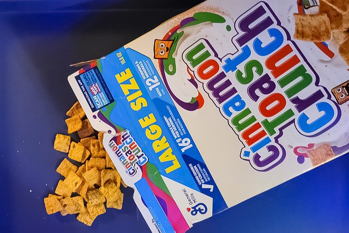 Spilled box of Cinnamon Toast Crunch cereal on a blue surface. Box displays "Large Size" and nutrition information.