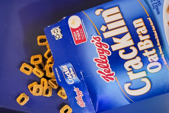 An open box of Kellogg's Cracklin' Oat Bran cereal on its side, with several cereal pieces spilled out onto a blue surface. The box features branding and nutritional information.