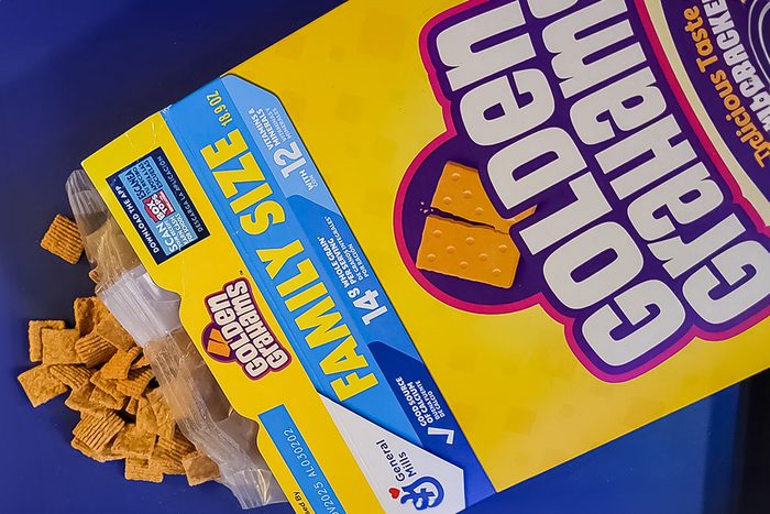 Box of Golden Grahams cereal titled "Family Size" on a blue surface, with several cereal pieces scattered beside it. The box is yellow with white and purple text and features images of cereal squares.