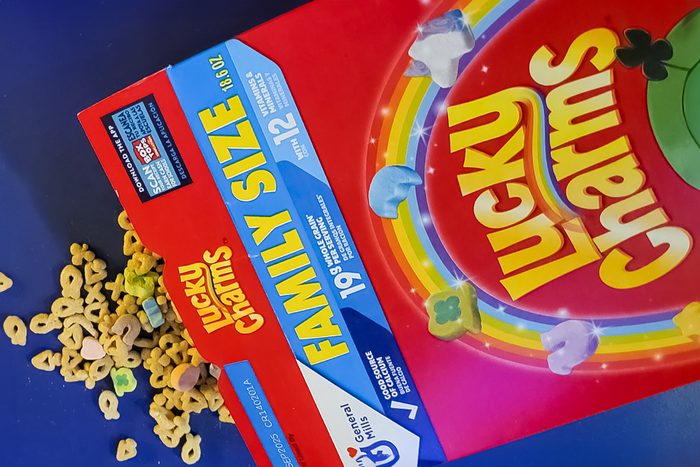 A box of Lucky Charms cereal labeled "Family Size" is tilted on a surface, spilling cereal pieces. The box features colorful packaging with the cereal's logo and images of marshmallow shapes.