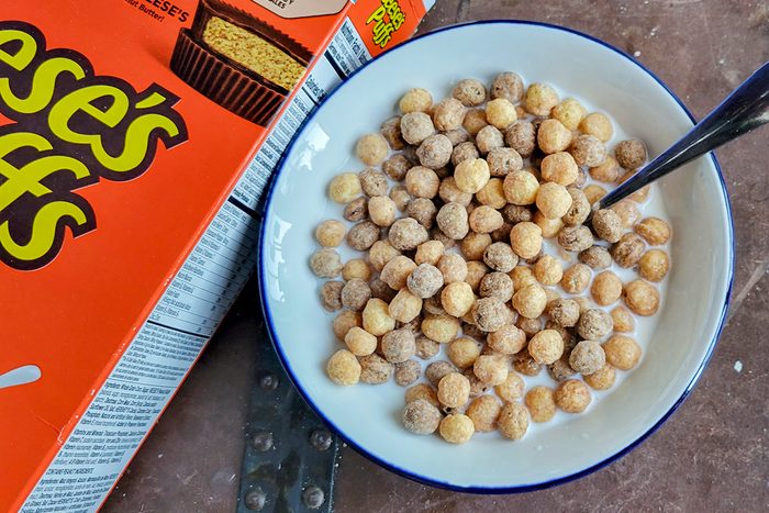 A bowl of cereal with milk and a spoon, featuring spherical, crunchy pieces. A partial view of a Reese's Puffs cereal box is visible next to the bowl. The surface is a textured brown.