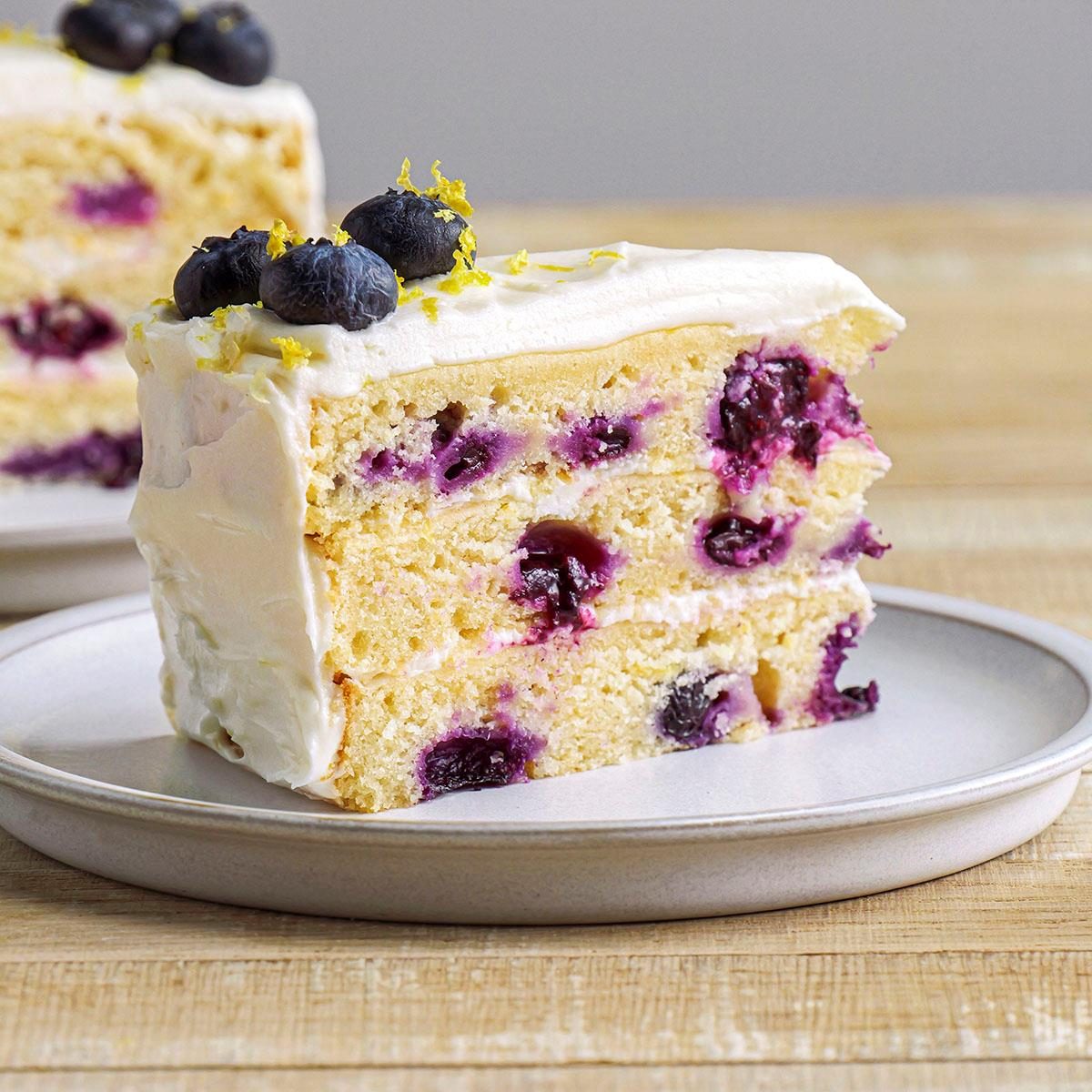 closeup view of lemon blueberry cake slices, the cake is topped with a creamy white frosting, which is garnished with a sprinkle of lemon zest and a few whole blueberries; resting on a round gray plate, this dessert is set against a rustic wooden background