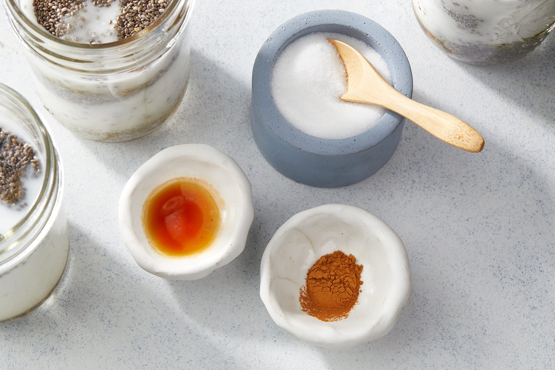 Overhead view of a white surface with ingredients in small bowls: chia seeds in glass jars, coarse salt with a wooden spoon, a bowl of sauce, and a bowl of ground cinnamon.