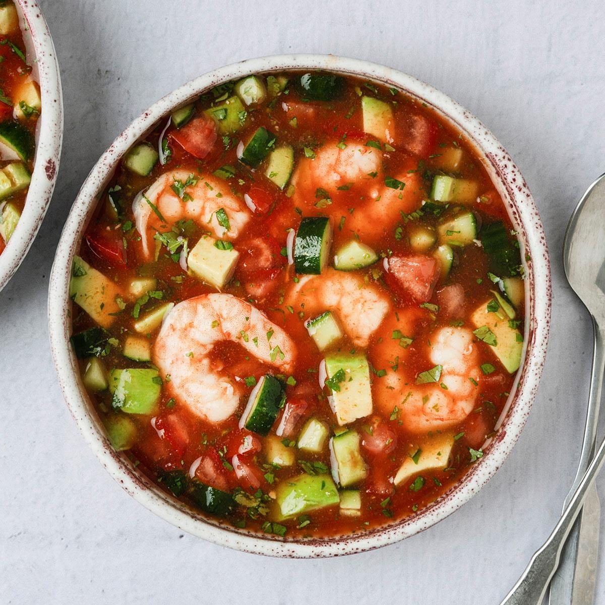 overhead shot of a dish called Shrimp Gazpacho; in two shallow bowls, next to the bowls are two shiny silver spoons, the background is a soft, light gray