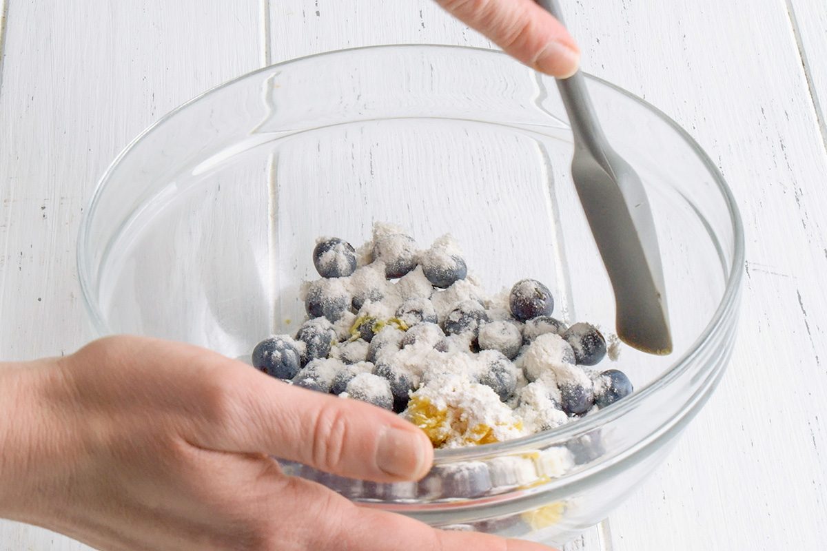 3/4th shot of Two hands are holding a clear glass bowl containing blueberries, flour or sugar, and small pieces of lemon zest; a gray spatula is being used to gently toss and combine these ingredients within the bowl; the bowl is positioned over a white wooden surface