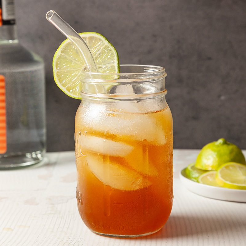 A mason jar filled with iced tea and ice cubes, garnished with a lime wheel on the rim and a glass straw. In the background, there’s a bottle, and a plate with sliced limes and lemons.